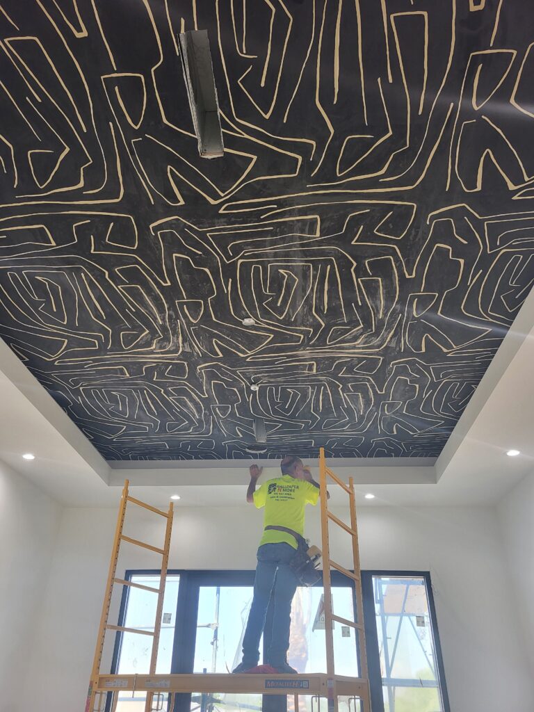 Worker standing on scaffolding installing a decorative black ceiling with intricate line patterns in a well-lit room.