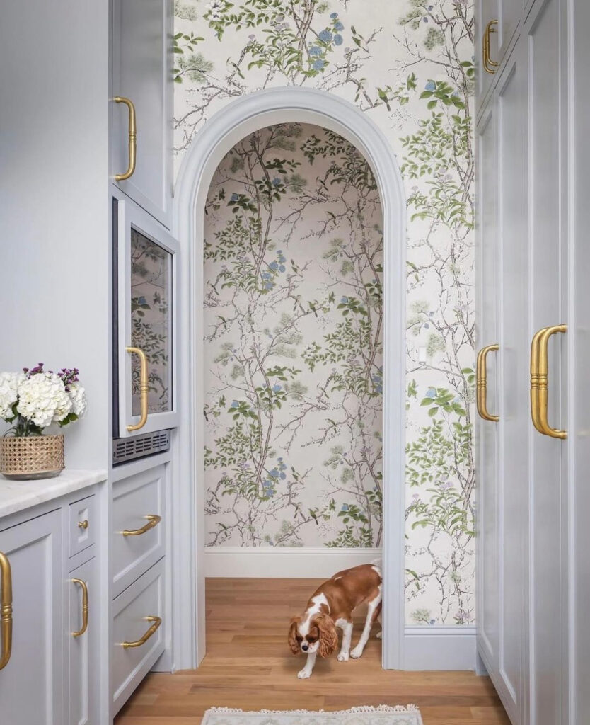 A small dog with brown and white fur stands in a hallway with floral wallpaper, wooden floor, and white cabinetry featuring gold handles.