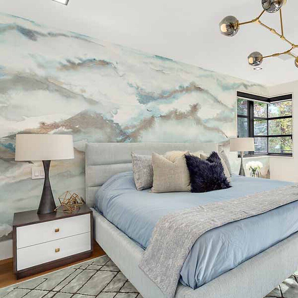 A modern bedroom with a blue upholstered bed, abstract wall art, geometric light fixture, two nightstands, and a patterned rug.