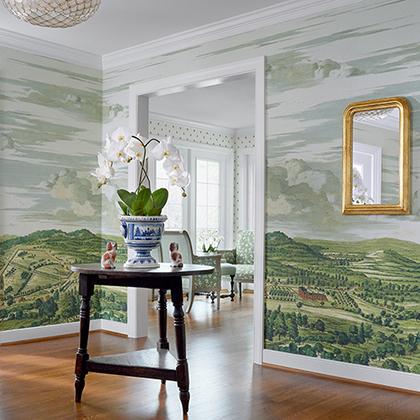 Living room with landscape mural, round wooden table with potted white orchids, gold-framed mirror, and hardwood floors.