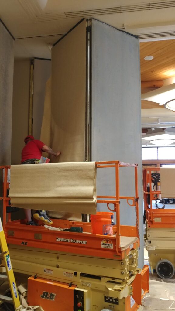 A worker wearing a red shirt and shorts operates a lift while installing or adjusting a large wall panel indoors.