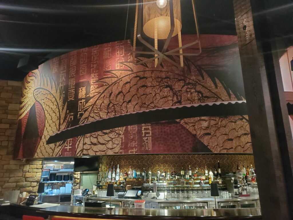 Bar area in a restaurant with a large mural featuring a dragon and Asian characters, bottles of alcohol displayed on the counter.