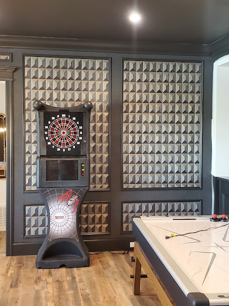 Room corner with a mounted electronic dartboard set, geometric-patterned walls, and a partial view of a game table and stool.