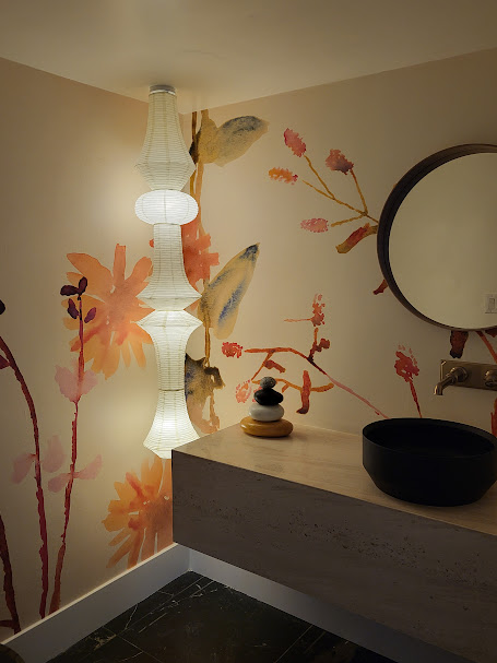 A bathroom with floral wall art, round mirror, rectangular sink, black basin, and vertical hanging white lamps.