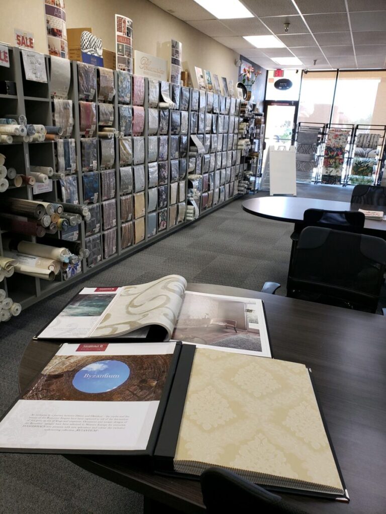 A fabric and wallpaper store with sample books on a table and shelves lined with various fabric rolls in the background.