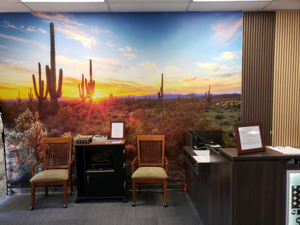 Office reception area with wall mural of a desert sunset behind cacti, wooden chairs, a side table with documents, and a reception desk.