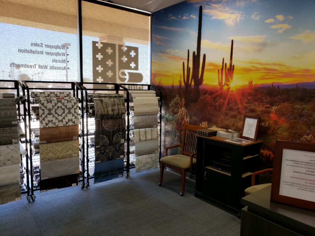 A showroom corner with tile samples on racks, a chair, brochures on a desk, and a scenic desert mural with cacti at sunset.