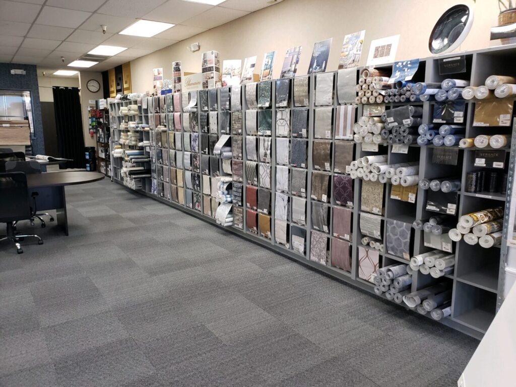 Shelves displaying various wallpaper rolls and samples in a store, with tables and chairs nearby for customer use.