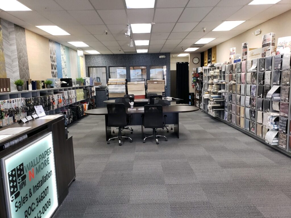 Interior of a wallpaper and flooring store with samples displayed on walls, a central table with chairs, and a sales counter on the left.