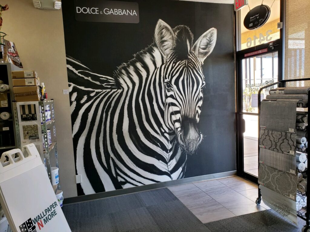 A store interior showcasing wallpaper with a large zebra mural. There are wallpaper samples and a "Wallpaper N' More" sign visible.