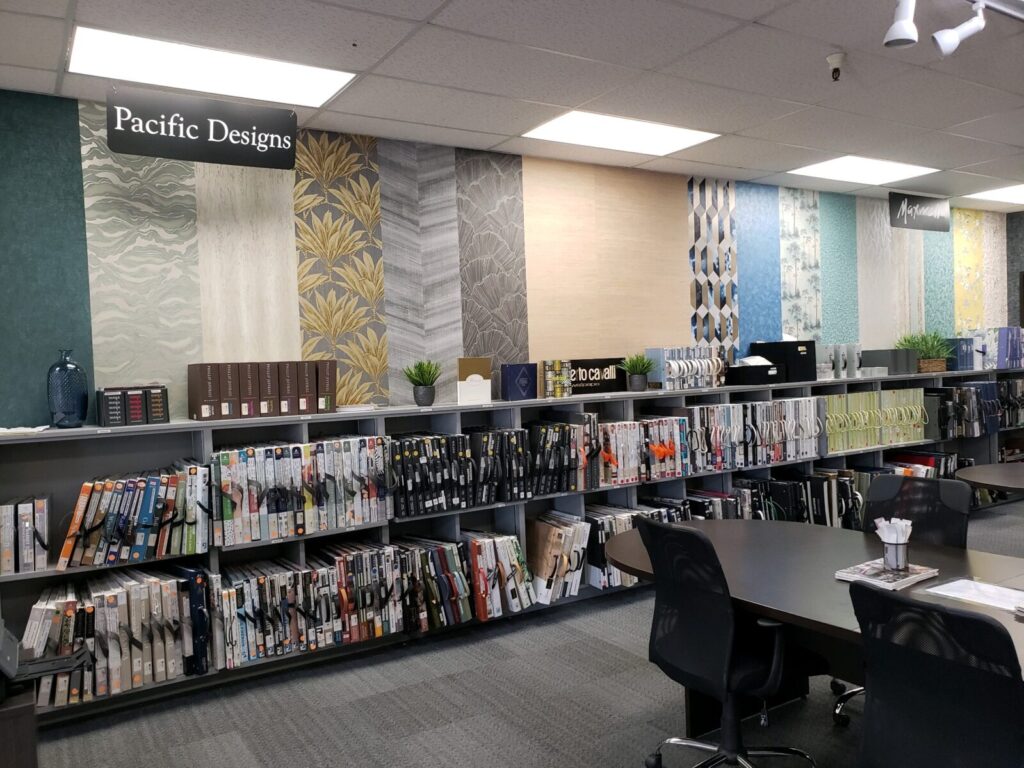 Interior of a design studio with samples of wallpaper and fabric swatches displayed on shelves and a large table with chairs in front.