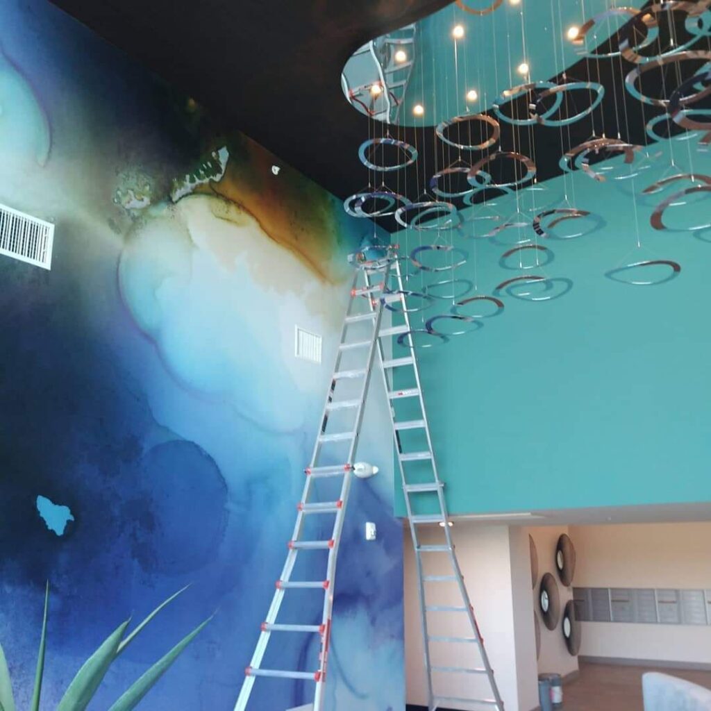 A room with a large colorful wall mural, a ladder leaning against the wall, and a modern hanging light fixture with circular elements.