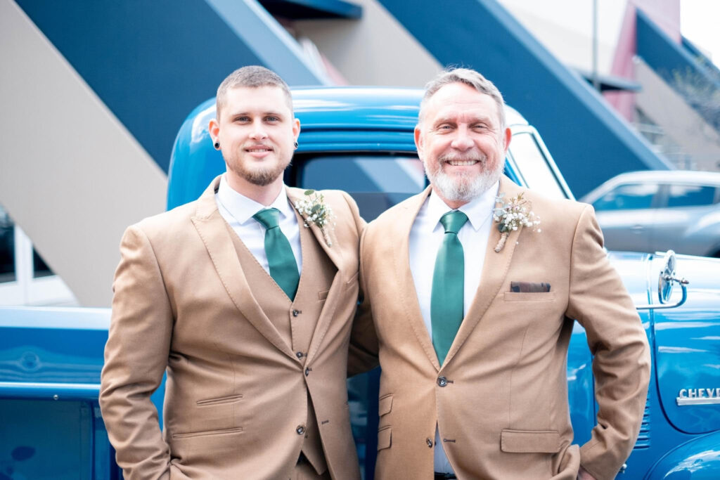 Two men wearing tan suits and green ties stand next to a blue vintage Chevrolet truck, smiling at the camera.